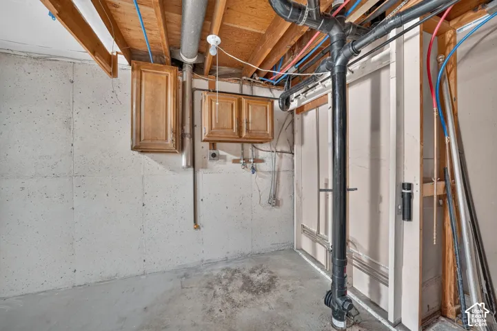 Unfinished utility space featuring concrete foundation walls, exposed wood-joist ceiling, wall-mounted wood-finish cabinetry, and exposed plumbing infrastructure
