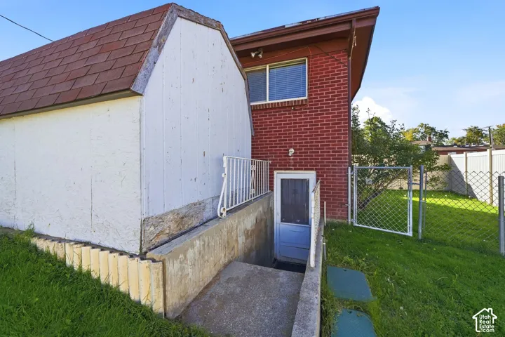 Red brick exterior siding, subterranean entrance with concrete staircase and metal handrail, detached shed with shingle roofing, and chain-link perimeter fencing
