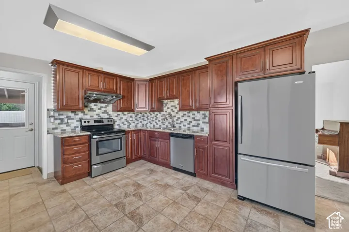 Kitchen featuring stained wood cabinetry, stone-tiled backsplash, stainless steel appliances, speckled stone-finish countertops, and ceramic tile flooring