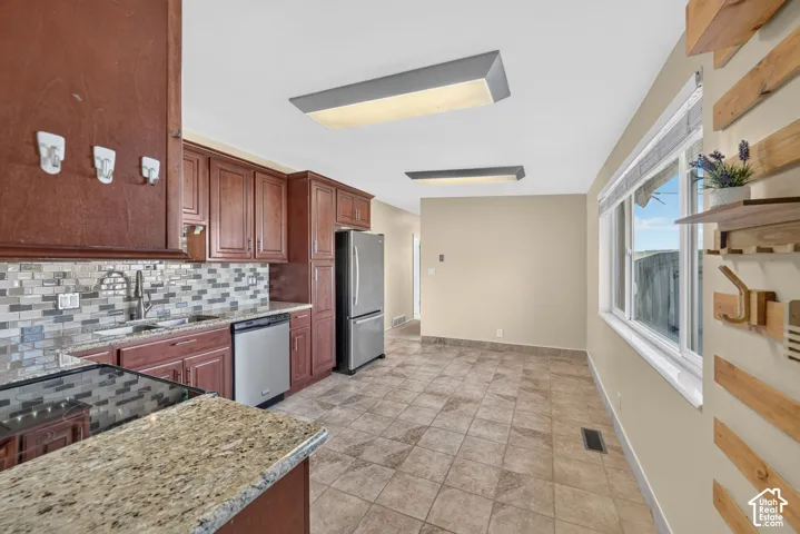 Kitchen featuring stained wood cabinetry, stone-finish countertops, a multi-tone tile backsplash, stainless steel appliances, and tile flooring