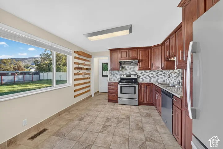 Kitchen featuring stained wood cabinetry, stone-patterned tile flooring, multi-tonal tile backsplash, stainless steel appliances, and a large window
