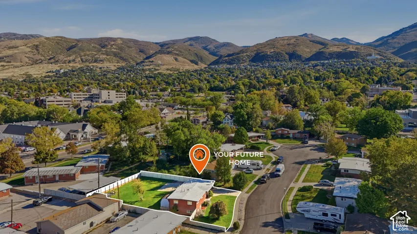 Aerial perspective featuring a brick exterior residence situated on a corner lot with a fenced backyard, mature trees, and a mountain backdrop