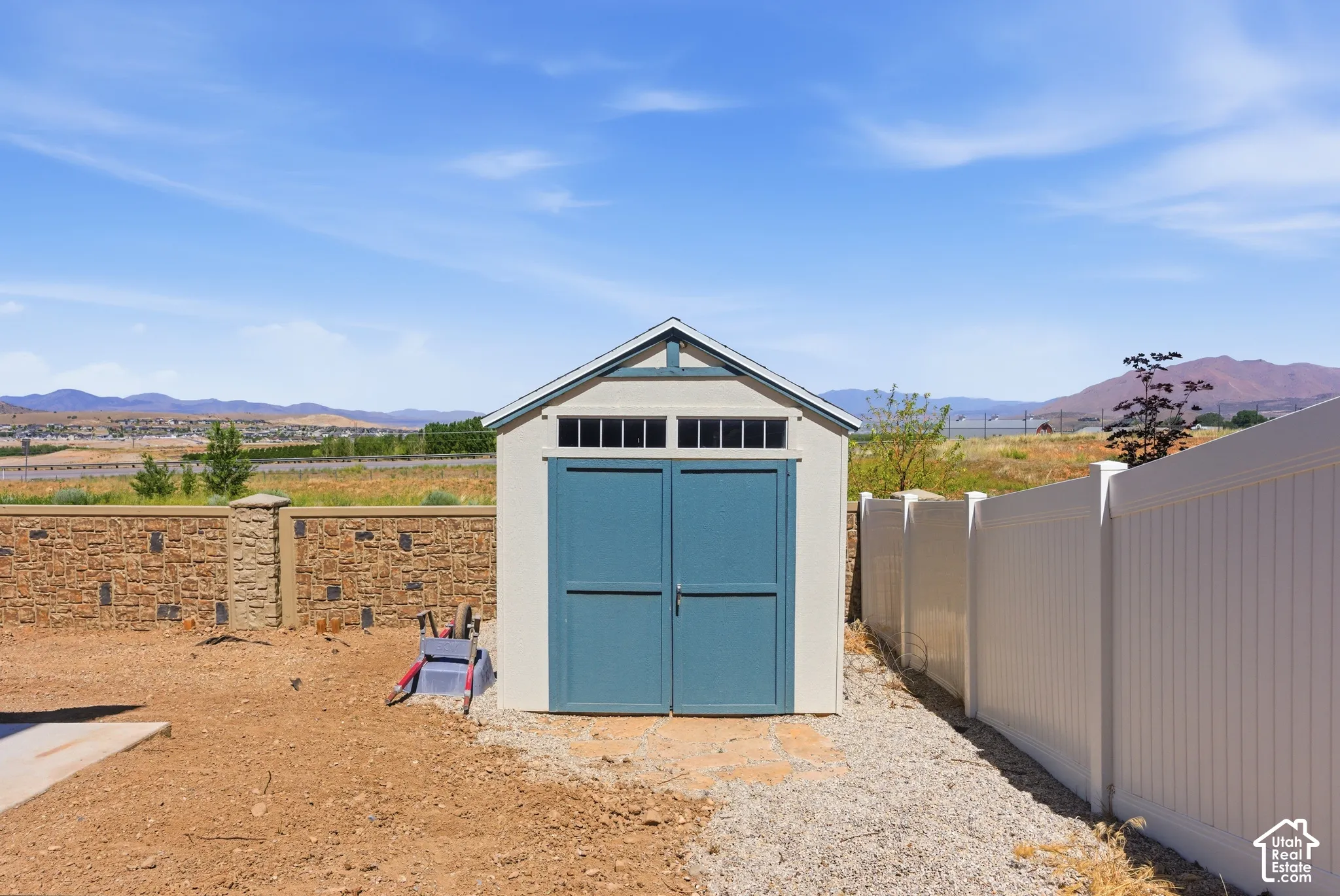 Detached storage structure featuring double doors, transom windows, and a gable roof, positioned within a fenced perimeter with stone-textured masonry and vinyl privacy fencing