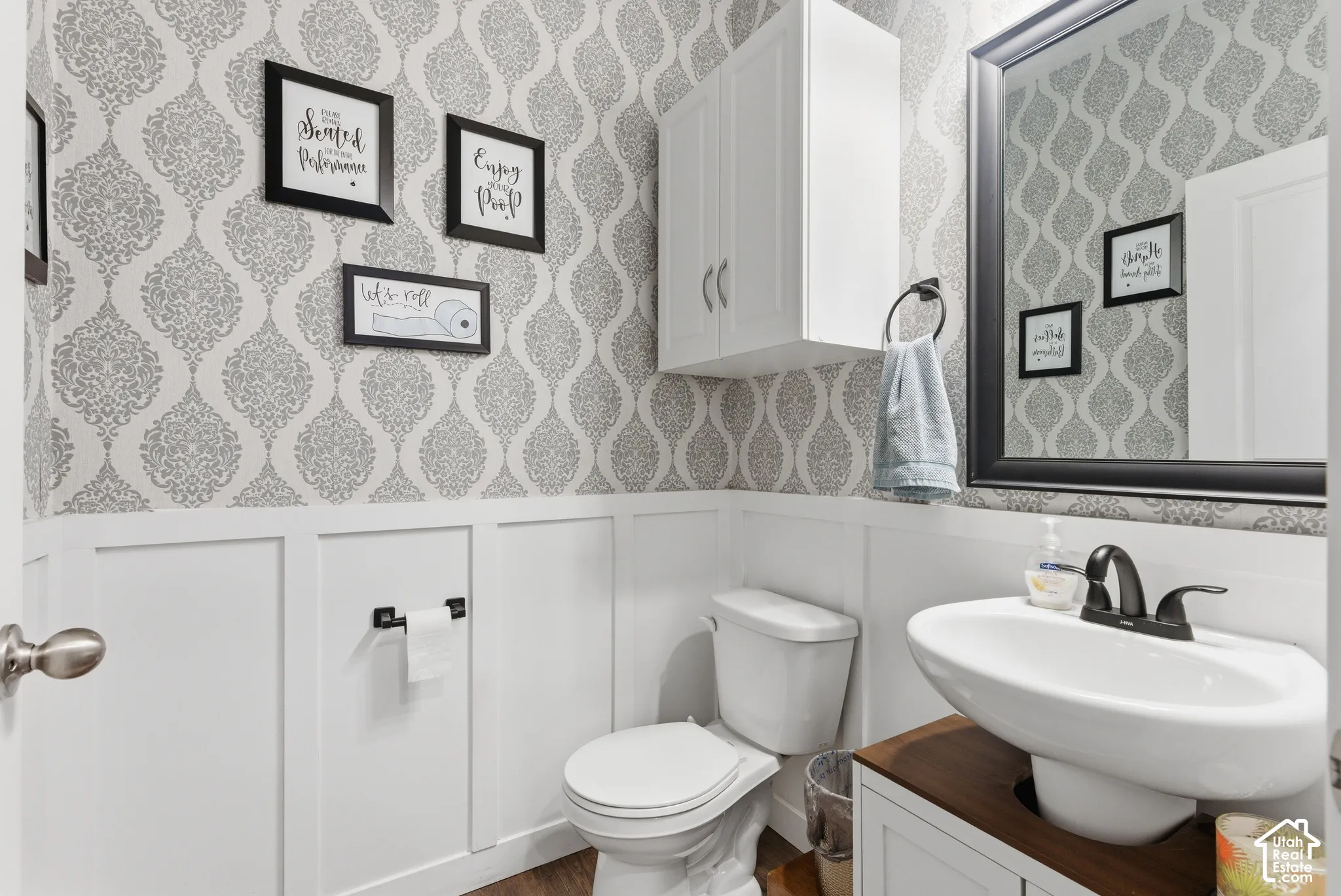 Powder room featuring patterned wallpaper, crisp white wainscoting, a pedestal-style sink basin set into a vanity, wall-mounted upper cabinetry, and a framed mirror with a matte black finish