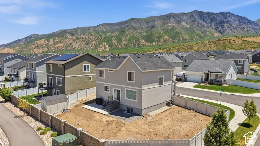 Two-story residence featuring a light-colored stucco and siding exterior, asphalt shingle roof, concrete patio, white vinyl perimeter fencing, and mountain backdrop