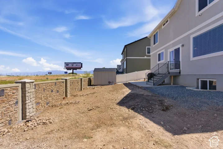 Rear exterior featuring a stone-patterned privacy wall, detached storage shed, metal staircase with railing, and gravel side yard area
