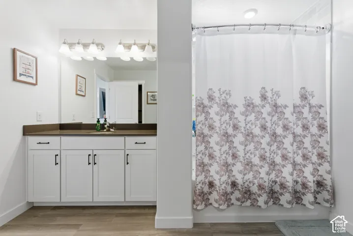 Bathroom vanity featuring white cabinetry, matte black hardware, a solid-surface countertop, and a wide wall-mounted mirror with multi-bulb light fixture, complemented by wood-finish flooring