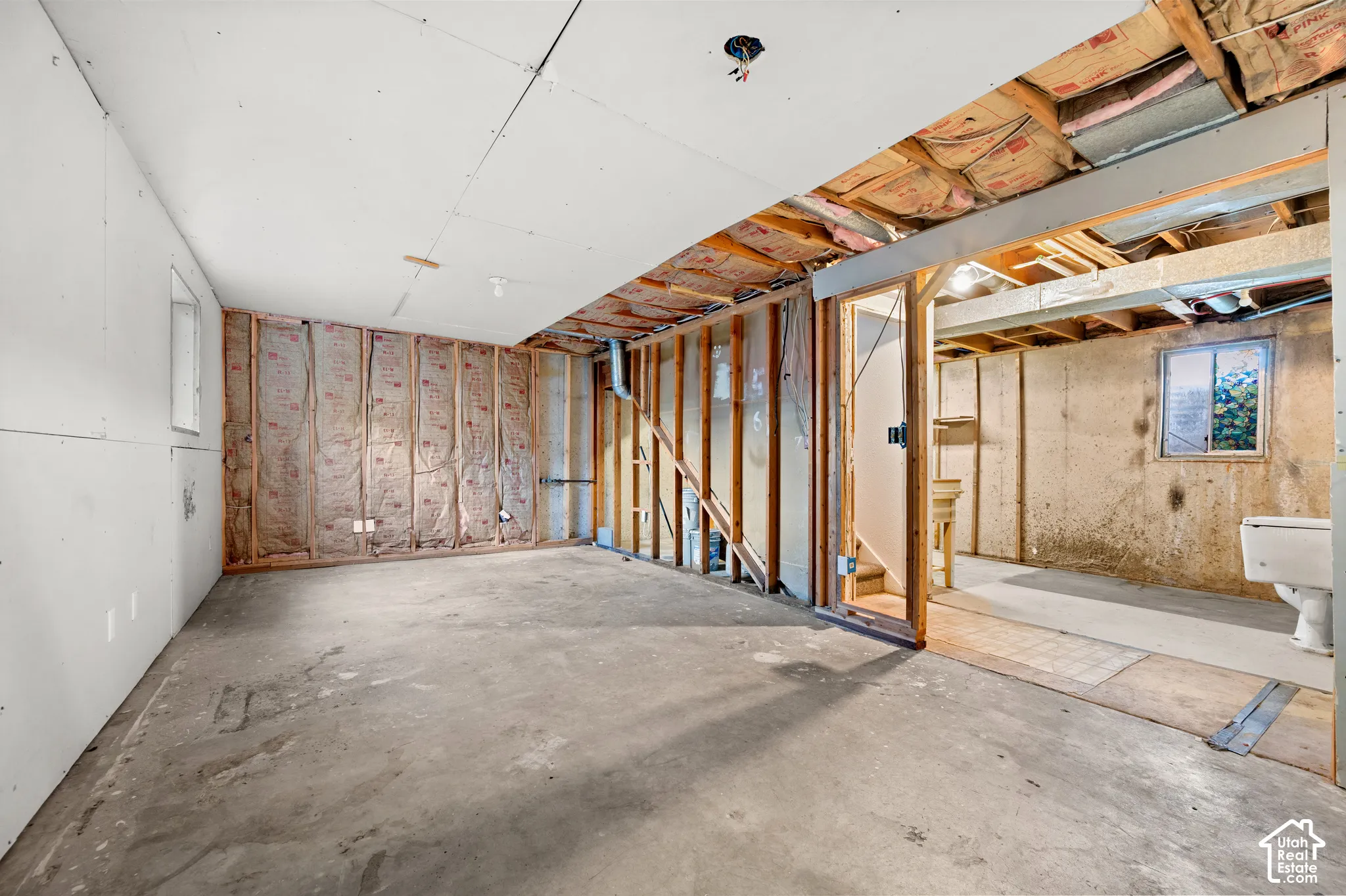 Unfinished basement area featuring concrete flooring, exposed wooden stud framing, exposed fiberglass insulation, and a single-pane window
