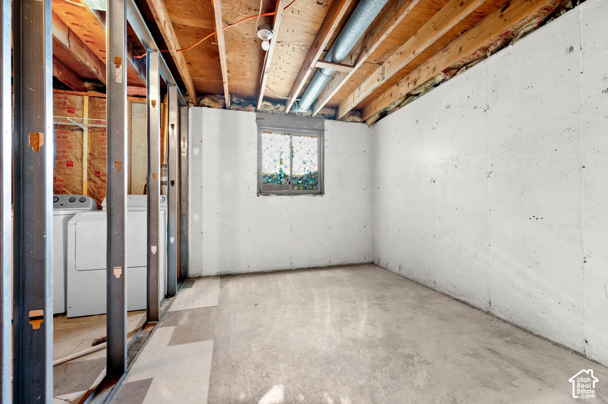 Unfinished lower-level space featuring concrete walls, exposed ceiling joists, metal stud framing, and a window