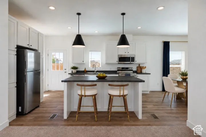 Kitchen featuring white shaker-style cabinetry, dark stone-finish countertops, stainless steel appliances, dual black pendant lights, and wood-finish flooring