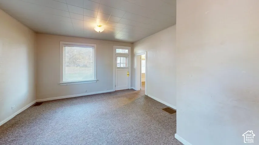 Neutral-toned interior featuring carpeted flooring, a grid-pattern ceiling, a single-light flush mount fixture, and a single-hung window with horizontal blinds