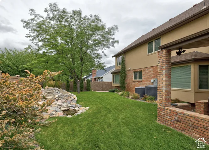 Backyard featuring a custom rock water feature, expansive lawn, brick-accented exterior, covered patio with brick columns, and privacy fencing