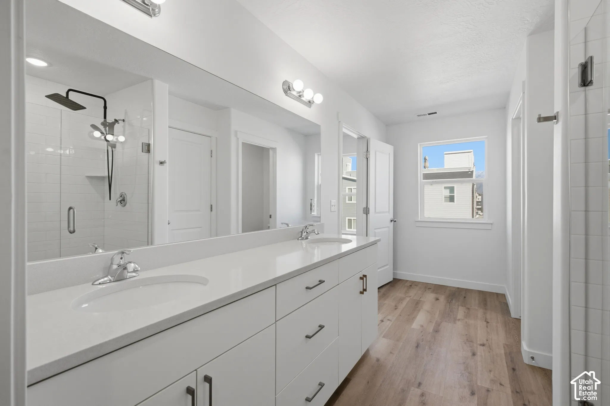 Double vanity featuring white cabinetry, a solid-surface countertop with dual undermount sinks, wood-finish flooring, and a walk-in shower with a matte black rainfall showerhead