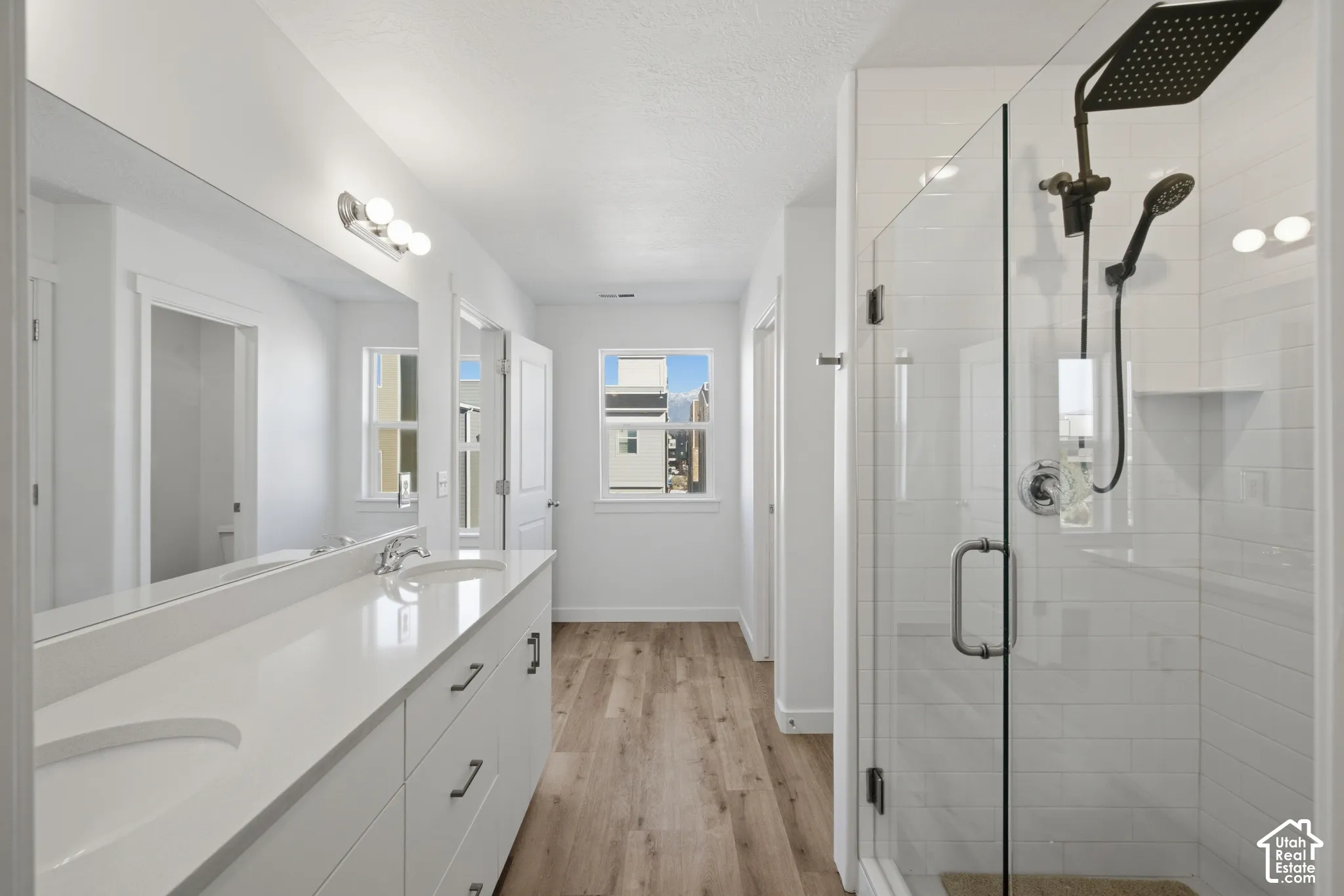 Dual-sink vanity with white cabinetry and solid-surface countertops, walk-in glass-enclosed shower featuring white subway-tile walls and a rainfall showerhead, and wood-finish flooring