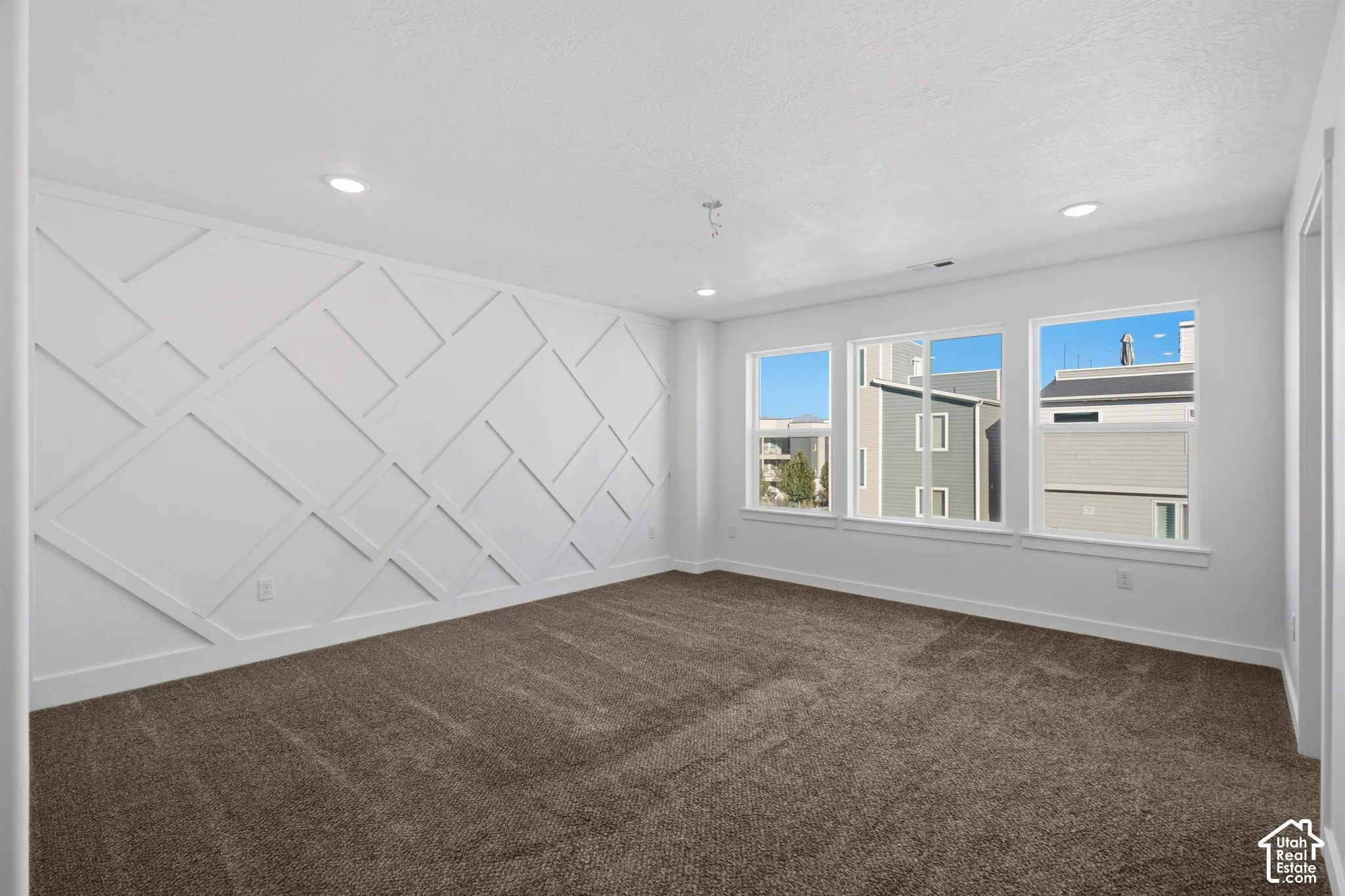 Room featuring a geometric wood wall accent, neutral carpeting, and multiple windows providing natural light, finished with recessed lighting