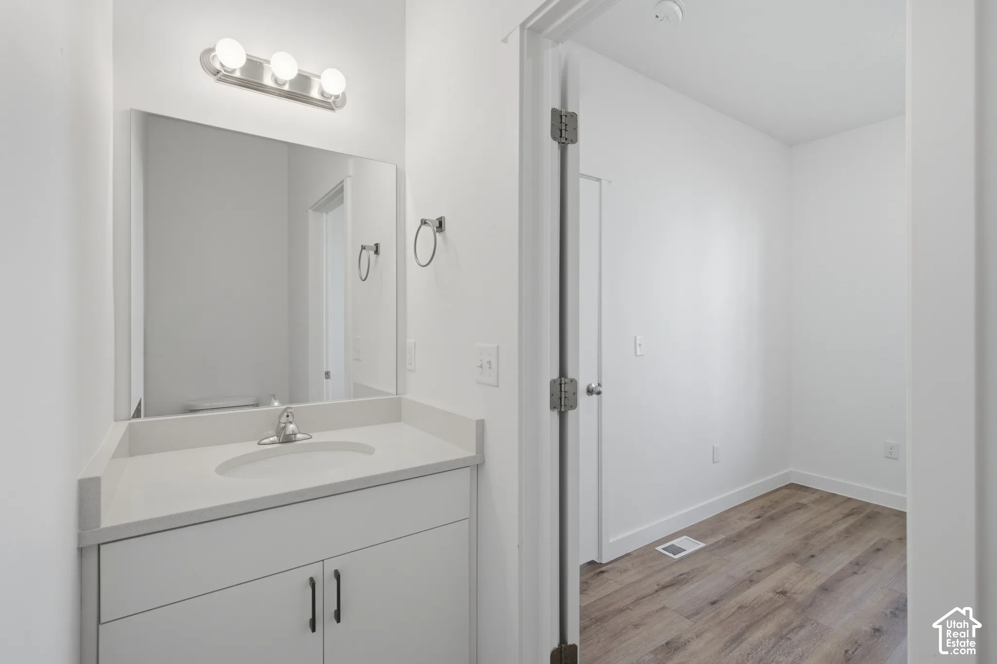 Vanity featuring a solid-surface countertop with an integrated basin, white shaker-style cabinetry with dark hardware, a rectangular wall-mounted mirror, triple-bulb light fixture, and wood-finish flooring