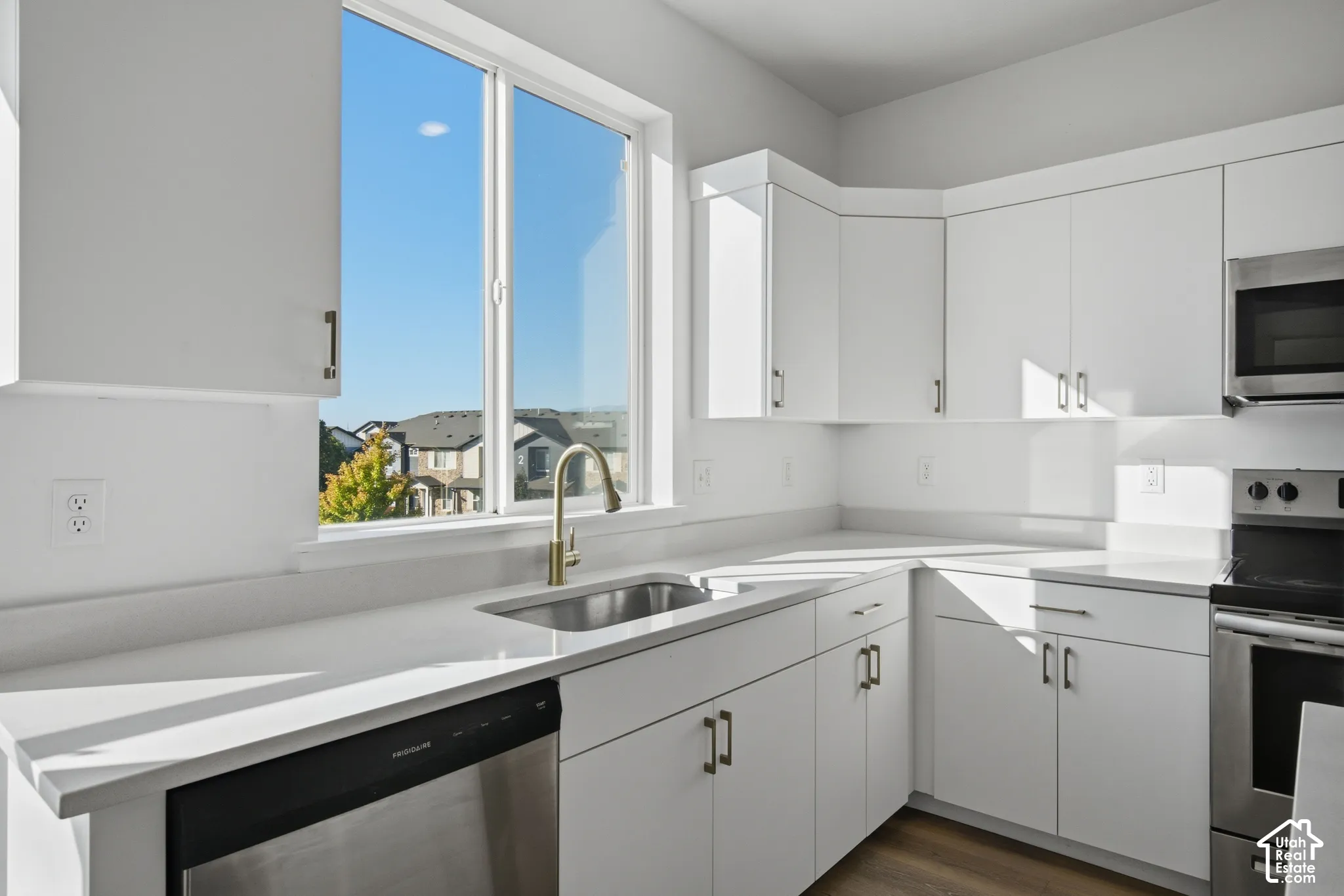 Kitchen featuring white shaker-style cabinetry, solid-surface countertops, stainless steel appliances, a deep undermount sink with a brass-finish faucet, and large window