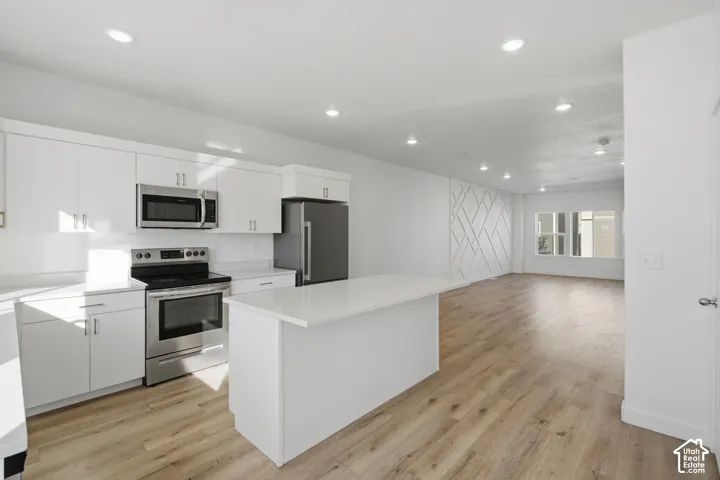 Kitchen featuring white cabinetry, an island with a light-toned countertop, stainless steel appliances, wood-finish flooring, and a decorative wall accent