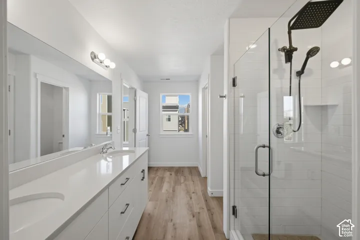 Dual-sink vanity with white cabinetry and solid-surface countertops, walk-in glass-enclosed shower featuring white subway-tile walls and a rainfall showerhead, and wood-finish flooring