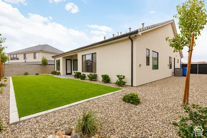 Stucco exterior with black window frames, featuring a fenced backyard, synthetic turf lawn, stone paver edging, and gravel ground cover