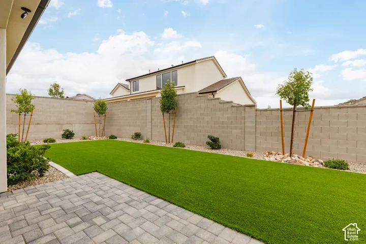 Backyard featuring a paved stone patio, artificial turf lawn, decorative stone perimeter, and concrete masonry wall