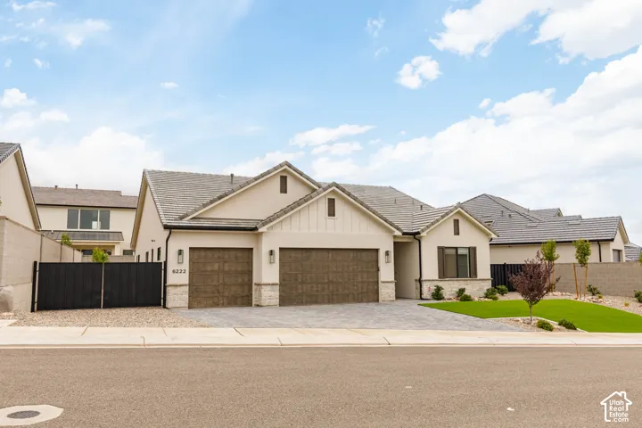 Exterior featuring a stucco facade with board-and-batten gable accents, multi-car garage with wood-finish doors, stone masonry base, concrete tile roofing, and paver driveway