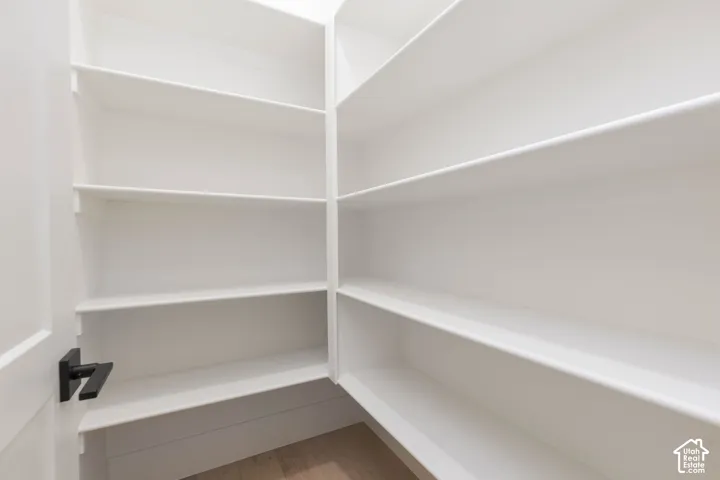 Walk-in storage space featuring floor-to-ceiling white shelving, neutral wood-finish flooring, and a matte black door handle