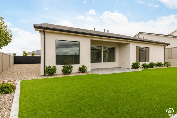 Stucco exterior with a recessed covered patio, synthetic turf lawn, stone-paved patio area, and perimeter masonry fencing