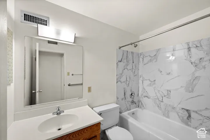 Bathroom featuring a wood-finish vanity, solid surface countertop with integrated sink, large wall-mounted mirror, and a tub with a stone-patterned tile surround