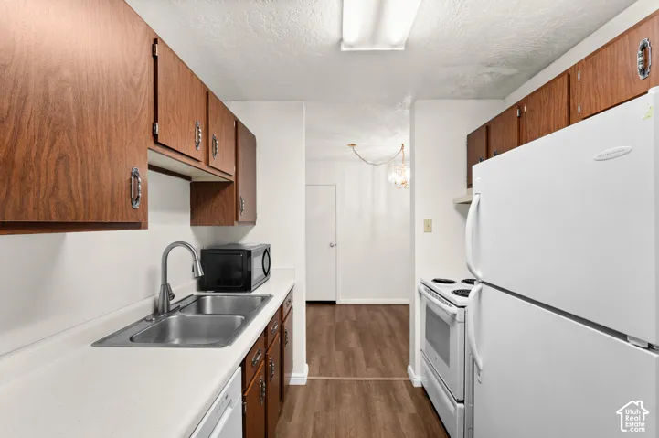Galley kitchen featuring wood-finish cabinetry, solid-surface countertops, dual-basin stainless steel sink with high-arc faucet, and white appliances