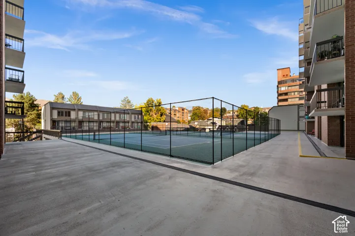 Fenced tennis court with hard-surface playing area, surrounded by concrete perimeter and multi-story structures