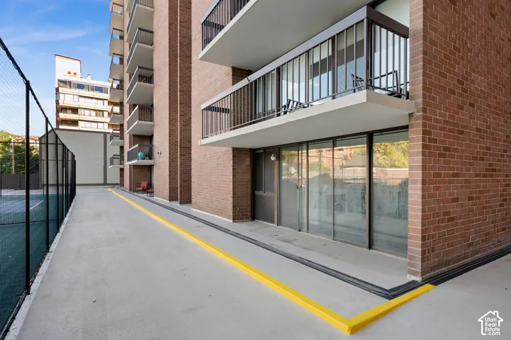 Red brick exterior featuring private balconies with metal railings, full-height sliding glass doors, and a paved outdoor court with a perimeter chain-link fence