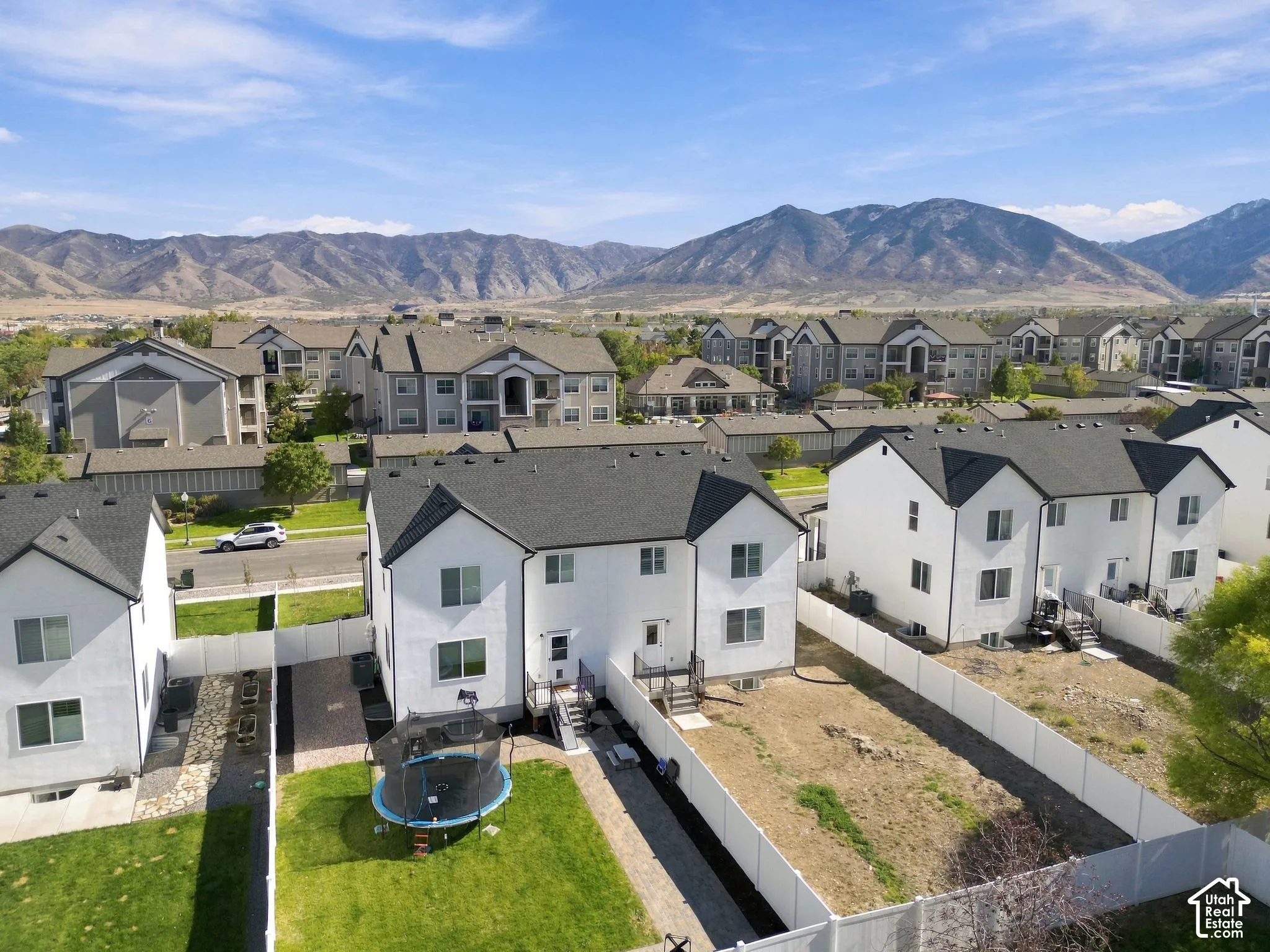 Aerial exterior featuring two-story residential structures with white stucco siding, dark shingled roofs, enclosed vinyl fencing, and mountain terrain backdrop