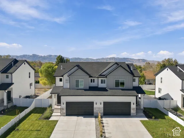 Two-story exterior featuring a mix of white stucco and grey vertical siding, dual-car garages, a dark shingle roof, stone veneer accents, and mountain backdrop