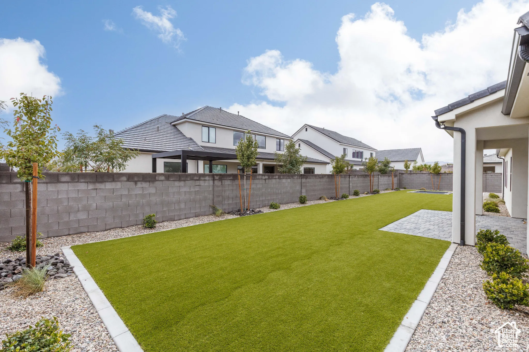Backyard featuring synthetic turf, concrete block perimeter wall, stone paver patio, decorative gravel borders, and young ornamental trees