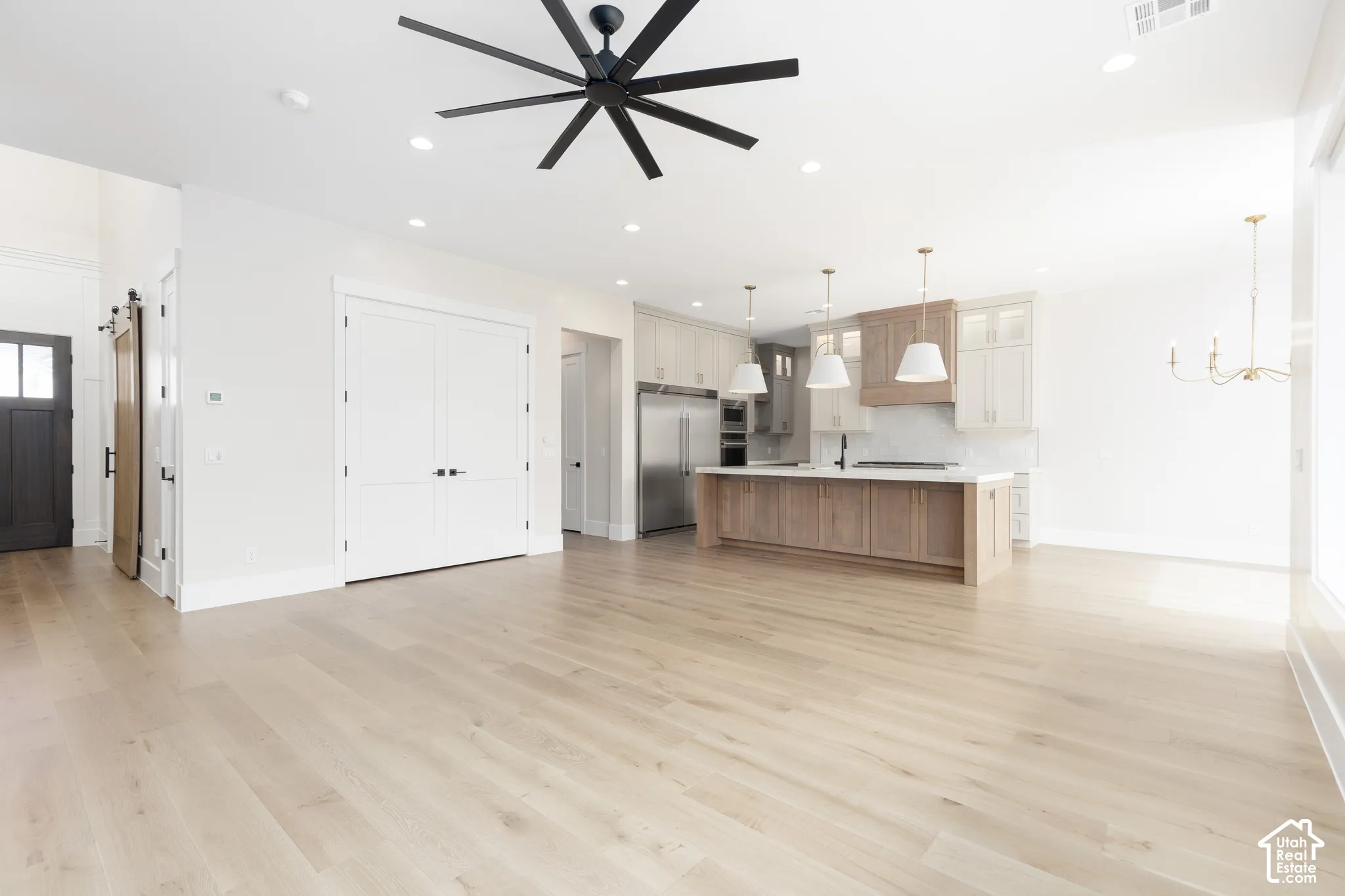 Open-concept interior featuring light wood-finish flooring, a kitchen island with stone countertops, stainless steel appliances, matte black ceiling fan, and recessed lighting