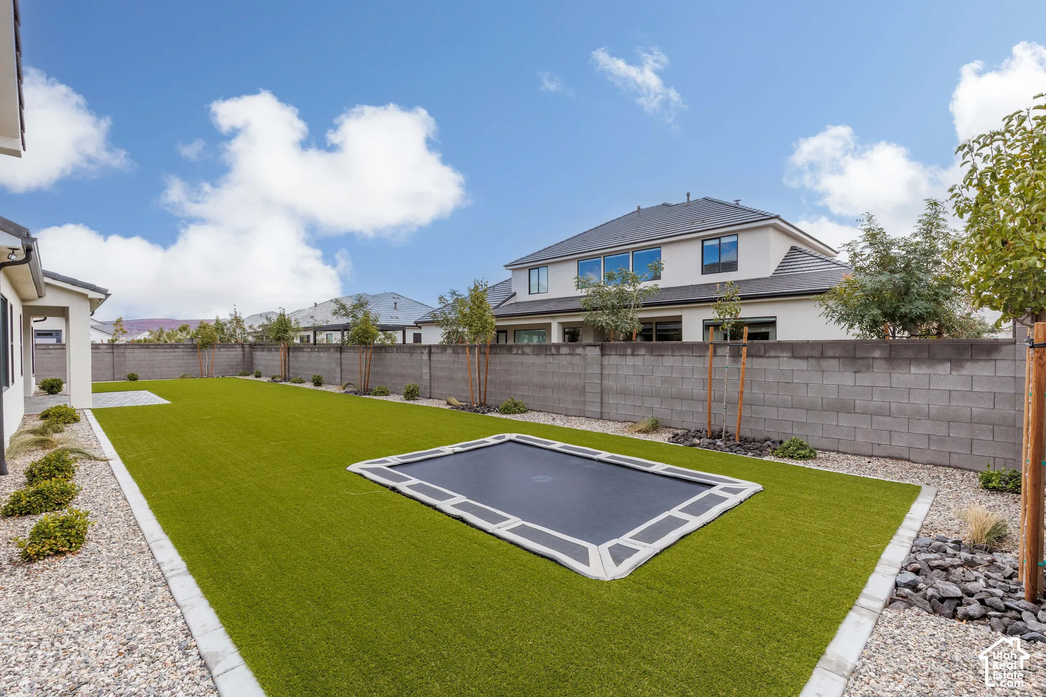 Backyard featuring synthetic turf, in-ground trampoline, concrete masonry unit perimeter wall, stone landscape bedding, and established young trees
