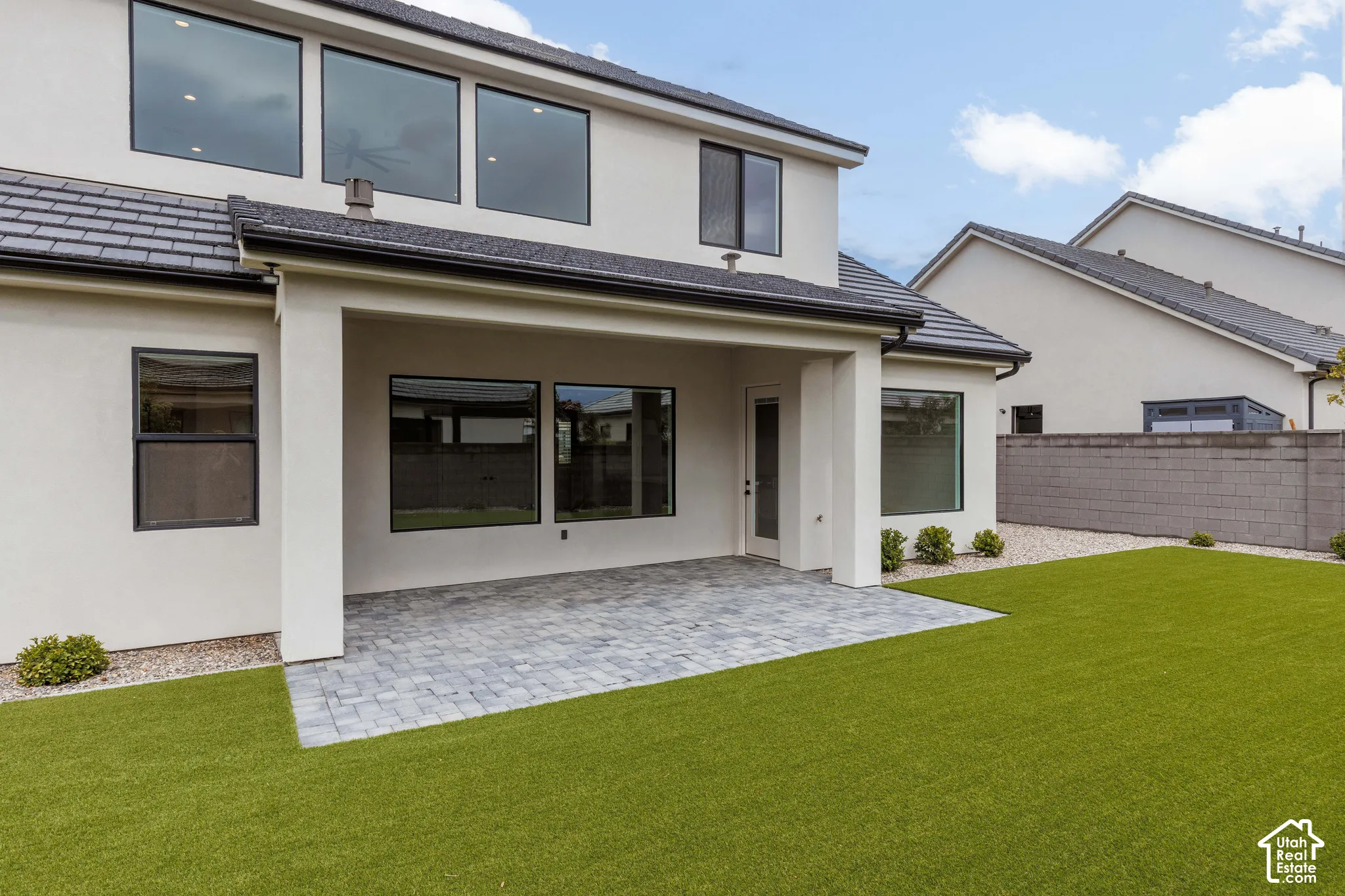 Stucco exterior featuring a covered paver patio, synthetic turf lawn, concrete block perimeter wall, and large-format multi-pane windows