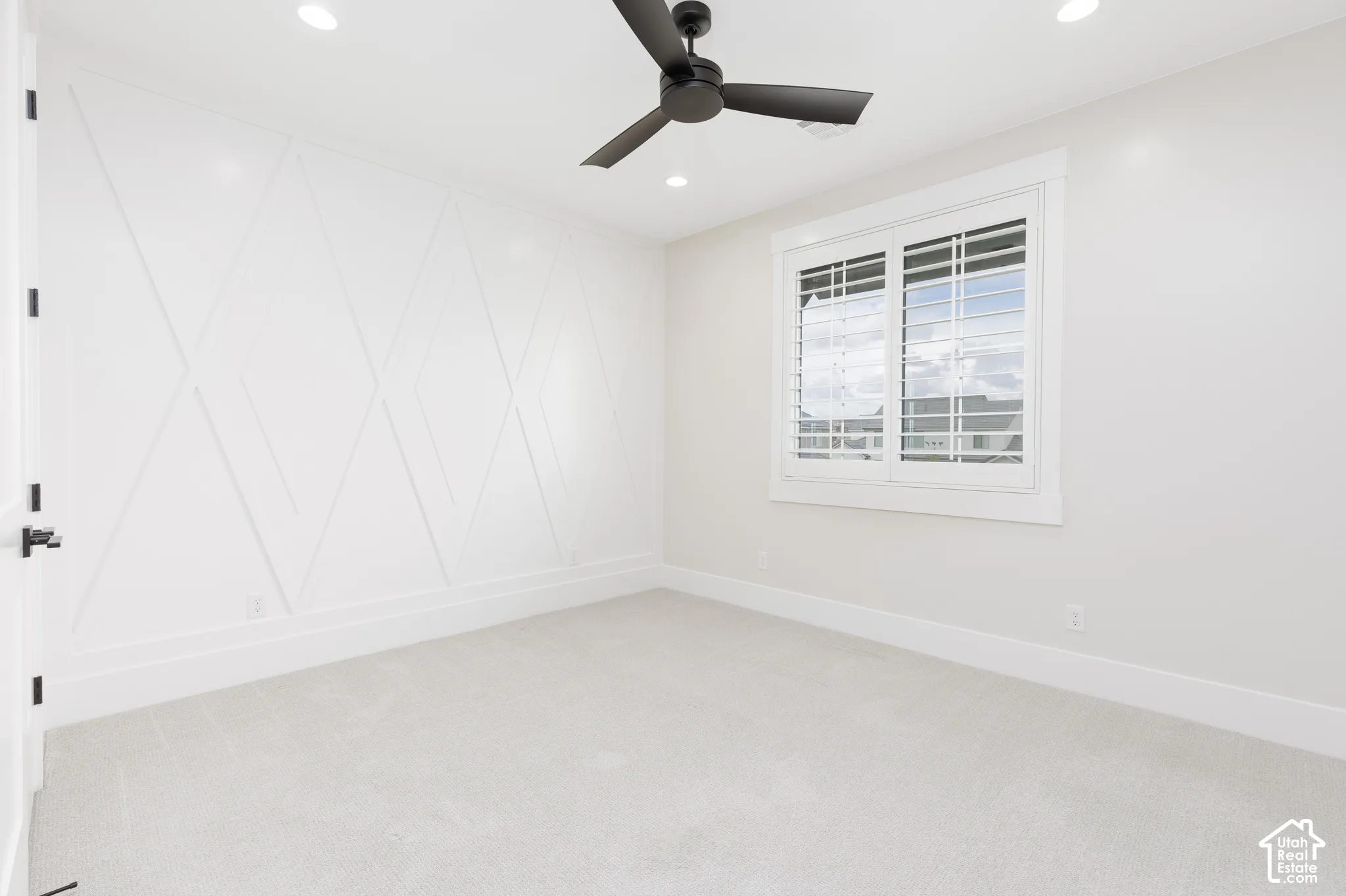Room featuring an accent wall with geometric millwork, neutral carpeting, a matte black ceiling fan, and window shutters