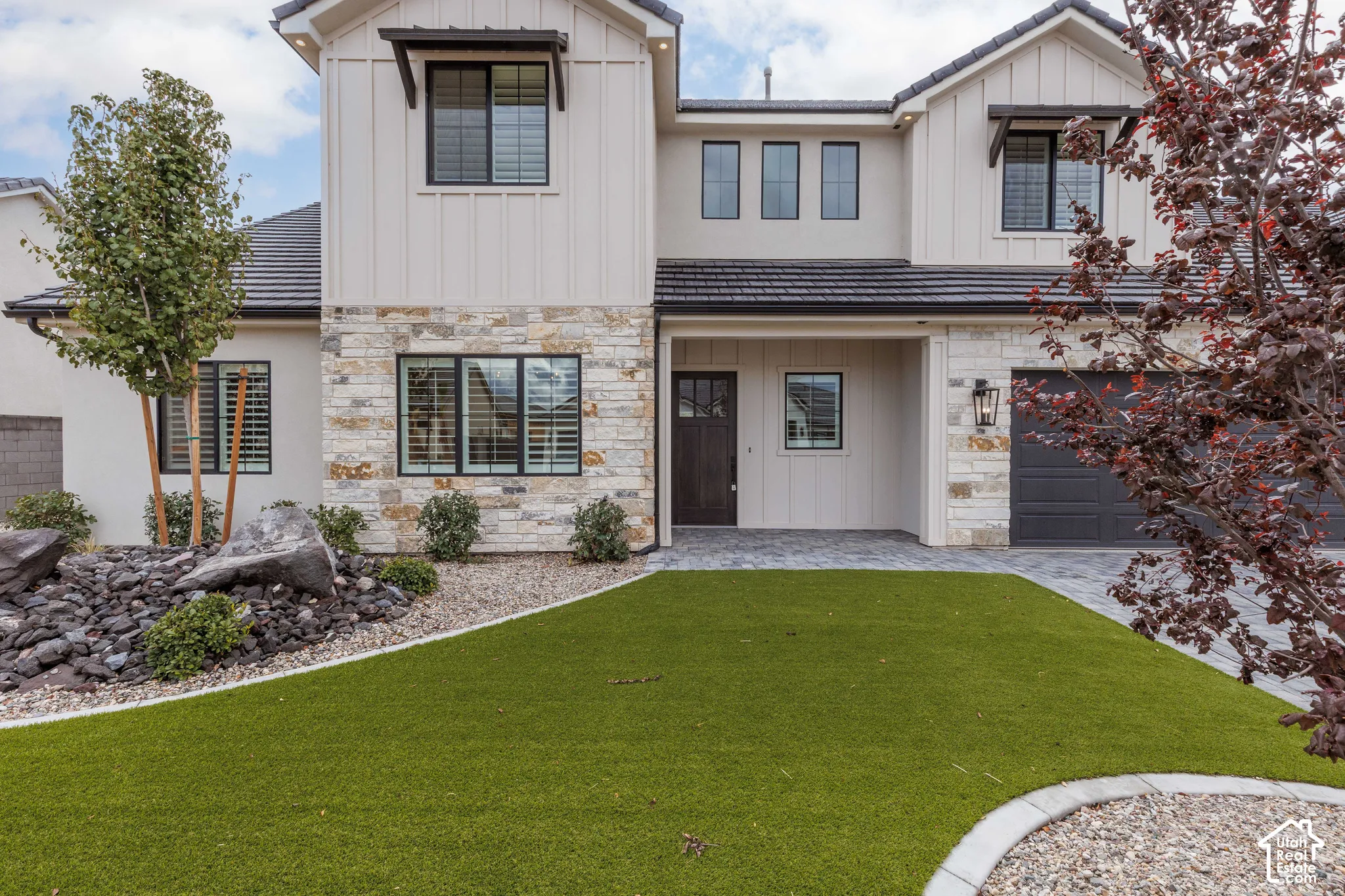 Contemporary two-story exterior featuring vertical board-and-batten siding, stacked stone masonry, a dark-toned tile roof, and a paved stone driveway