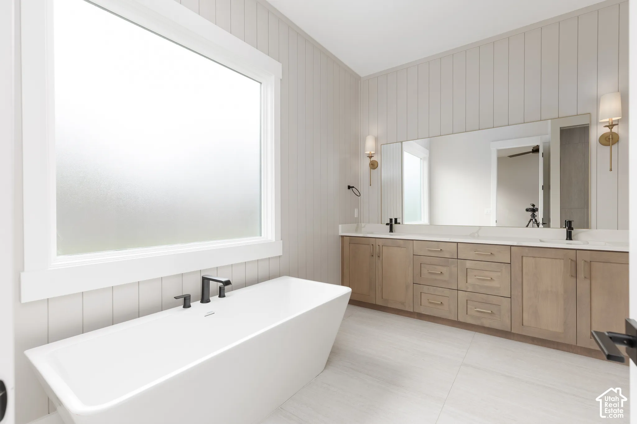 Freestanding soaking tub with matte black deck-mounted faucet, double vanity featuring light wood cabinetry, white stone countertop, large wall-to-wall mirror, and vertical shiplap wall paneling