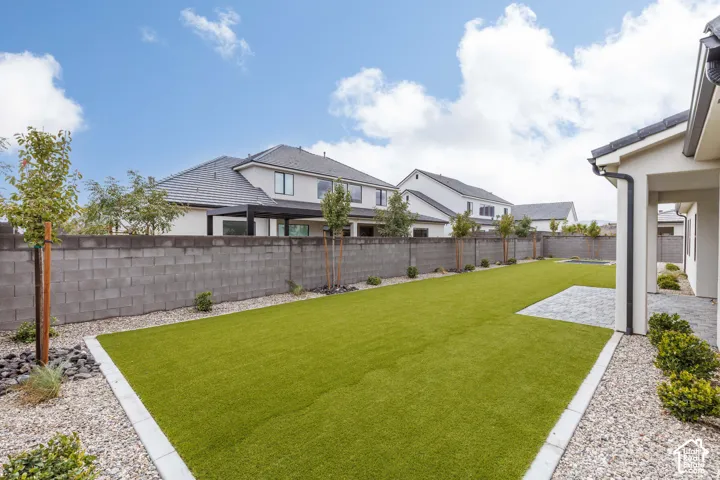 Backyard featuring synthetic turf, concrete block perimeter wall, stone paver patio, decorative gravel borders, and young ornamental trees