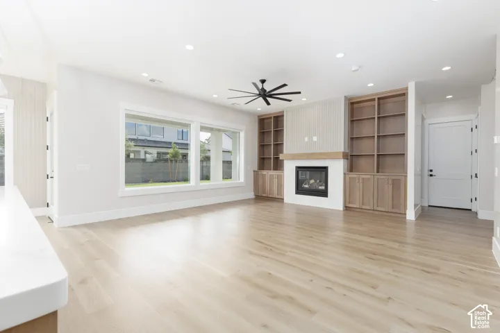 Living area featuring wood-finish flooring, a fireplace with a wood mantel, built-in cabinetry with shelving, large picture windows, and a ceiling fan