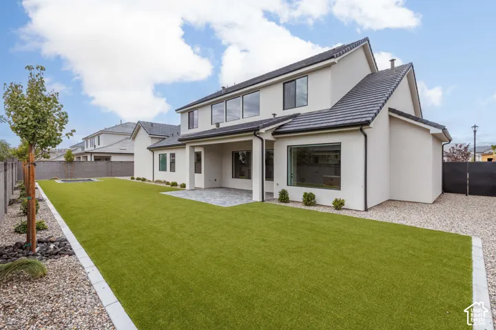 Stucco exterior featuring a covered stone-paved patio, synthetic turf lawn, concrete curbing, and a perimeter masonry wall