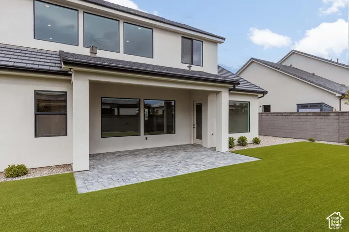 Stucco exterior featuring a covered paver patio, synthetic turf lawn, concrete block perimeter wall, and large-format multi-pane windows