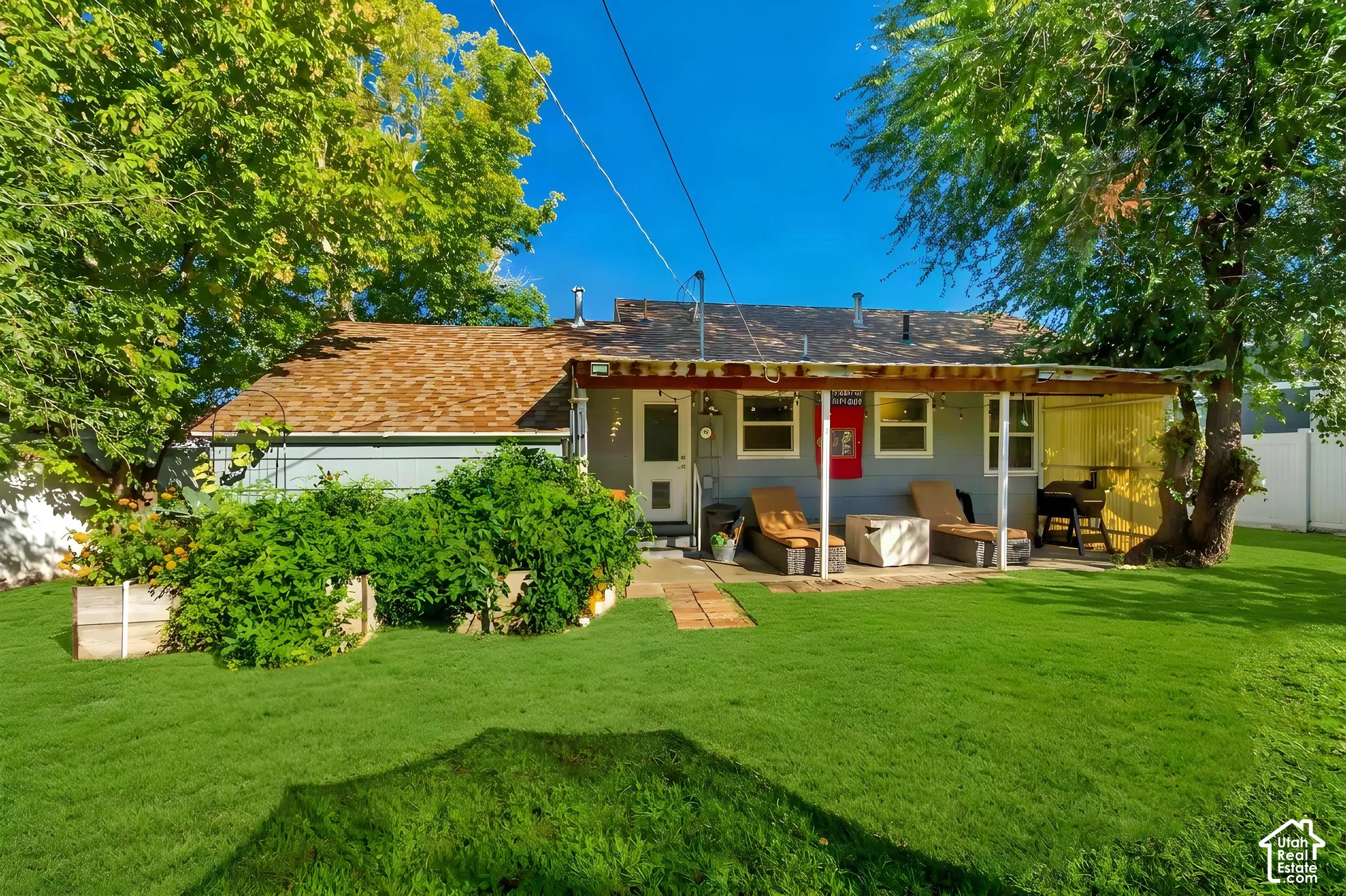 Rear exterior featuring a covered patio, paved hardscaping, mature trees, and raised garden beds