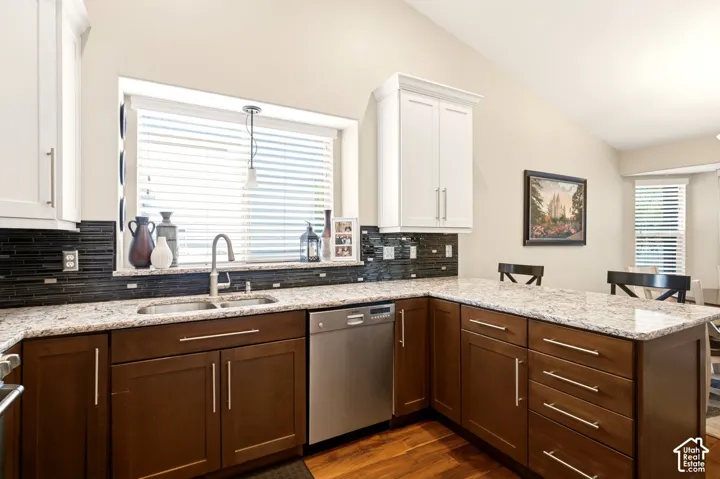 Kitchen featuring stone-finish countertops, dark wood-finish cabinetry with contrasting white upper cabinets, black linear tile backsplash, stainless steel dishwasher, and wood-finish flooring