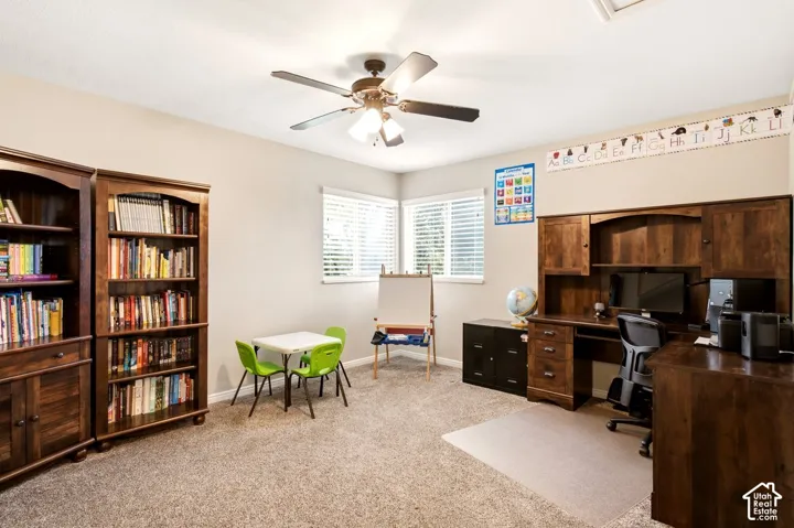Neutral-toned interior room featuring wall-to-wall carpeting, dual windows with blinds, a multi-blade ceiling fan with integrated lighting, and extensive built-in wood cabinetry including shelving and a workspace desk