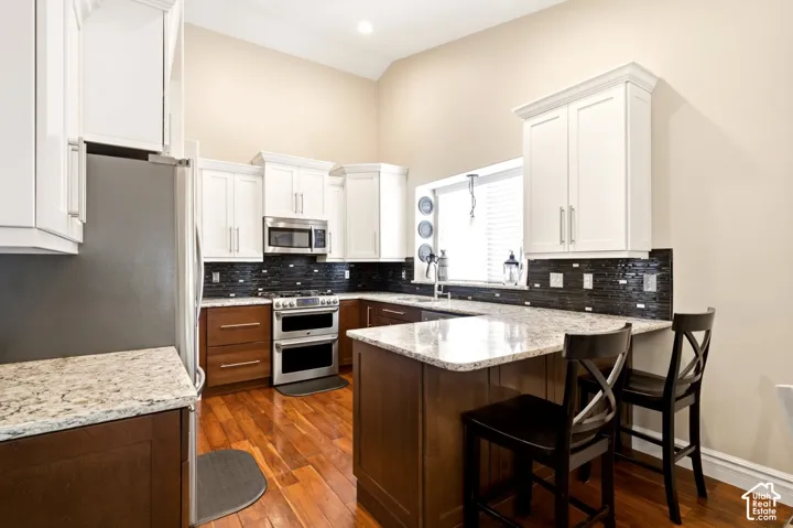 Kitchen featuring two-tone cabinetry, stone countertops, dark tile backsplash, stainless steel appliances, and wood-finish flooring under vaulted ceilings