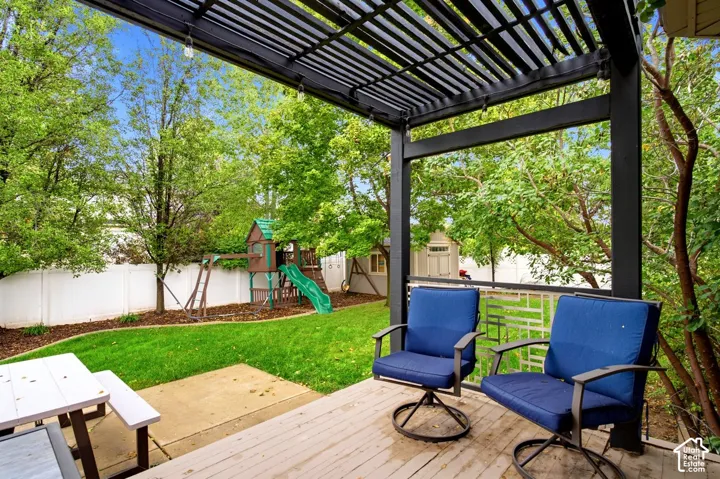 Covered deck featuring a black metal pergola, decorative metal privacy screen, wood-surface decking, manicured lawn, and white vinyl perimeter fencing